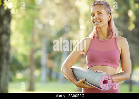 Portrait von glücklichen jungen Frau im rosa Top holding Trainingsmatte und lächeln Sie im Park zu trainieren Stockfoto
