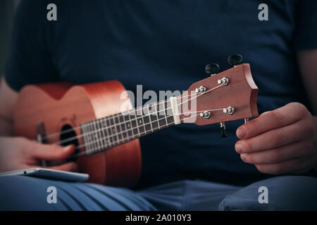 Der Mann in der blauen Kleidung Tunes die Saiten der Ukulele, mit der Anwendung auf dem Telefon, der auf seinem Knie liegt. Stockfoto