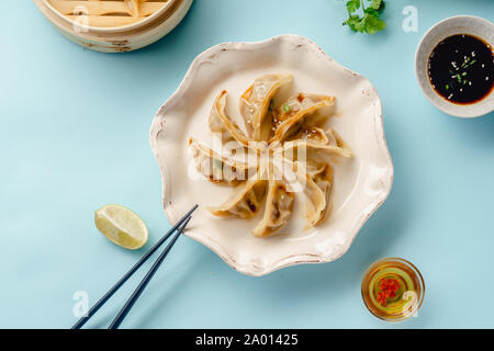 Gyoza-teigtaschen mit Ente in Bambus Steamer gegart, serviert auf einem Teller mit Sojasoße, Sesam und Koriander. Ansicht von oben, blauer Hintergrund Stockfoto