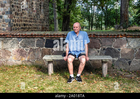 Älterer Mann sitzt auf der Bank vor der Mauer aus Stein von Pinnow Dorfkirche, Uckermark, Brandenburg, Deutschland Stockfoto