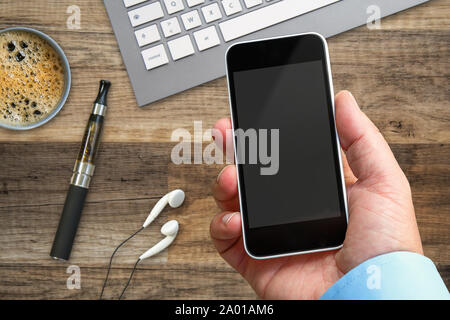 Smartphone in der Hand bei der Arbeit mit einem leeren Bereich um ein benutzerdefiniertes Bild einfügen. Viele Objekte auf dem hölzernen Tisch platziert Stockfoto