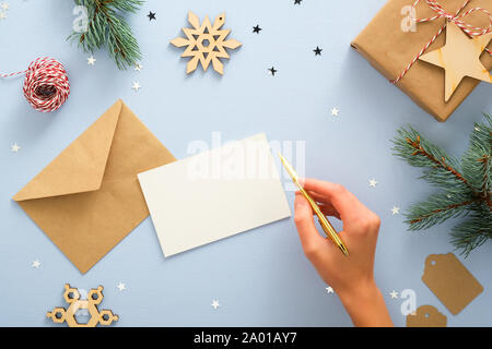 Weibliche Hand schreiben Brief an Santa Claus über blauen Hintergrund mit festlichen Weihnachtsschmuck, Konfetti, Fir Tree Branches, Schneeflocken, Geschenkbox. Stockfoto