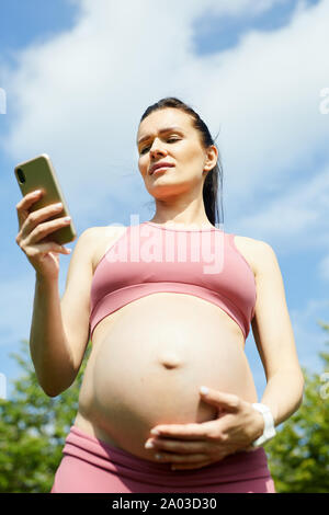 Schöne schwangere Frau in rosa Top halten ihr Handy in der Hand und kommunizieren online beim Stehen im Freien Stockfoto