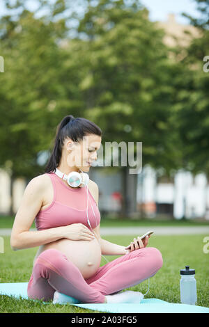 Junge schwangere Frau in rosa Kleidung sitzen auf trainingsmatte und über Handy beim Sport Training im Park Stockfoto