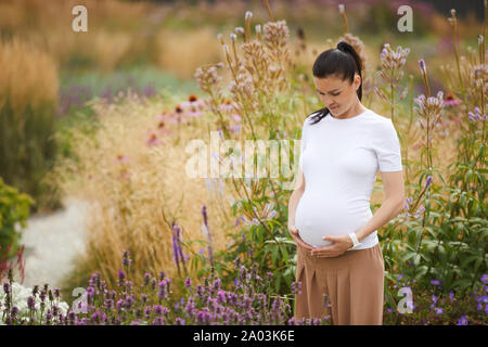 Schöne Frau mit dunklen Haaren in Freizeitkleidung ihrer schwangeren umfassende Bauch beim Stehen im Freien mit Blume Bereich im Hintergrund Stockfoto