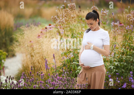 Nette junge schwangere Frau auf ihren Bauch, während in der Nähe des Bereich der Blumen im Freien stehend Stockfoto