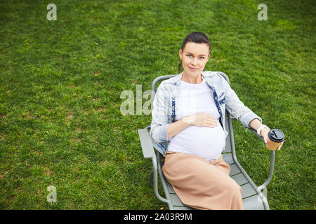 Portrait von jungen schwangeren Frau auf Stuhl und trinkt Kaffee im Freien im Sommer Tag Stockfoto