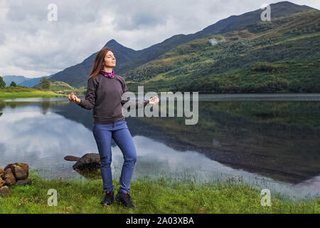 Junge Frau Tourist in der Nähe der sehr ruhigen und friedlichen See mit ihren Händen in meditativen Position und mit geschlossenen Augen Genießen der Natur stehende Stockfoto