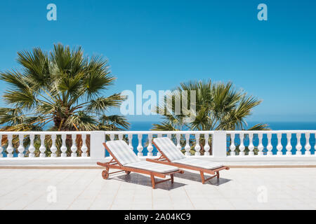 Sonnenliegen auf der Terrasse mit Blick aufs Meer und Palmen im Hintergrund Stockfoto