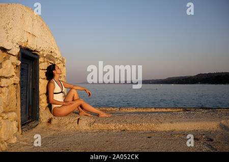 Junge schöne Frau in einem Badeanzug und Sonnenbrille sitzt auf einem Pier in Griechenland bei Sonnenuntergang. Mädchen genießt die Sonne Stockfoto