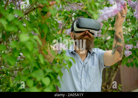 Mann mit VR-Brille wandern in üppigen grünen Garten, moderne Technologien Konzept. Hipster mit tätowierten Arm holding Zweig von Blooming Tree. Bärtige Stockfoto