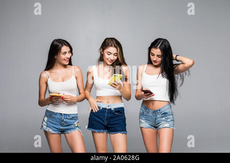 Drei hübsche junge Mädchen legere Kleidung nach oben auf der Suche und Nutzung von Mobiltelefonen über grauer Hintergrund Stockfoto