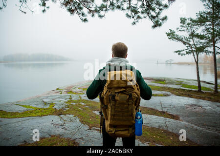 Reifer mann Finnland erkunden im Herbst, in Nebel. Wanderer mit grossen Rucksack auf bemoosten Felsen. Skandinavische Landschaft mit misty Meer und Stockfoto