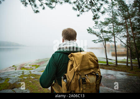 Reifer mann Finnland erkunden im Herbst, in Nebel. Wanderer mit grossen Rucksack auf bemoosten Felsen. Skandinavische Landschaft mit misty Meer und Stockfoto