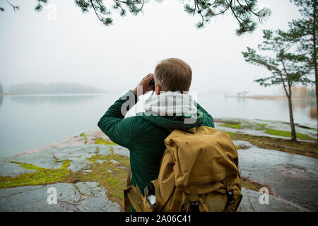 Reifer mann Finnland erkunden im Herbst, in Nebel Blick durch ein Fernglas. Wanderer mit grossen Rucksack auf bemoosten Felsen. Skandinavische Landschaft Stockfoto