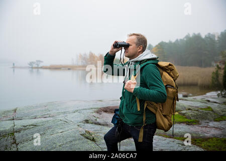 Reifer mann Finnland erkunden im Herbst, in Nebel Blick durch ein Fernglas. Wanderer mit grossen Rucksack auf bemoosten Felsen. Skandinavische Landschaft Stockfoto