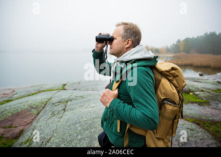 Reifer mann Finnland erkunden im Herbst, in Nebel Blick durch ein Fernglas. Wanderer mit grossen Rucksack auf bemoosten Felsen. Skandinavische Landschaft Stockfoto