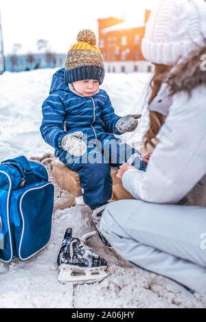 Kleiner Junge von 3,5 Jahre alt, Winter auf Stadt Eislaufbahn, sitzt auf der Bank, Frau s Mutter ihr Kind s Kleidung, setzt auf Schuhe, Rest am Wochenende Stockfoto