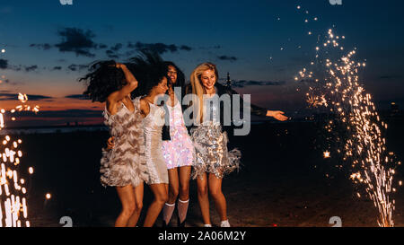 Gruppe von Freundinnen gerne tanzen zwischen Feuerwerk in der Nacht. Junge stilvolle Frauen feiern bei Sonnenuntergang. Stockfoto