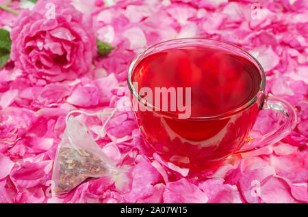 Tasse heißen Blumentee auf einem Hintergrund von Rosenblättern, liegt neben einem Teebeutel. Selektiver Fokus. Stockfoto