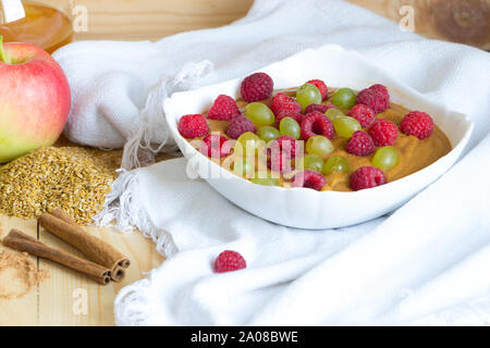 Vegan Flachs Porridge mit Himbeeren und Trauben, Rosinen in weißen Schüssel auf Leinen. Leinsamen, Zimtstangen, gereift Apfel und Honig im Glas Stockfoto