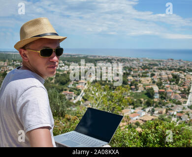 Ein Freiberufler Mann in einem Hut und Brille schaut die Kamera beim Sitzen auf einem Berg, mit einer schönen Landschaft, einer Stadt am Meer, Arbeiten in den Plädoyers Stockfoto