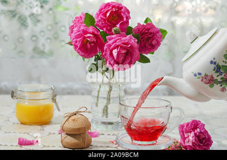 Heiße Tasse flower Tee wird aus den Wasserkocher gegossen, neben Cookies, Honig und Rosen. Selektive konzentrieren. Stockfoto