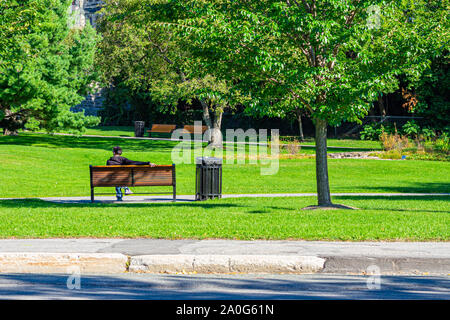 Ein Mann lehnt sich zurück und streckt einen Arm über den Rücken von einer Parkbank, weg von der Straße (und Kamera). Stockfoto