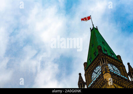 Das Zifferblatt der Uhr, Dach und Flagge auf der Oberseite des Peace Tower in Kanadas Parlamentsgebäude wird von unten gegen einen blauen bewölkten Himmel gesehen. Stockfoto