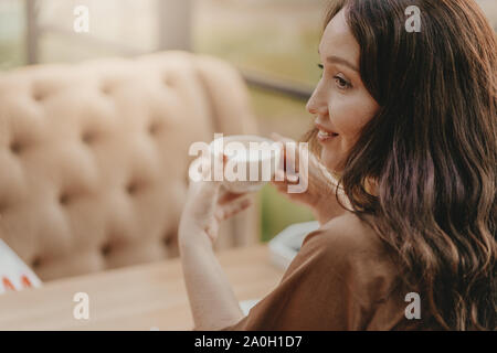 Hübsche brünette Frau mit langen lockigen Haar am Fenster sitzen im Cafe bei einer Tasse Kaffee in der Hand Stockfoto