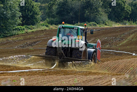 Arc von Gülle in der Sonne gefangen wird, es wird auf einen Drei-tage-Feld durch ein langes Rohr aus der Farm auf einem John Deere Traktor. Stockfoto