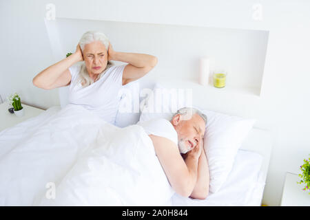 Entspannen, ausruhen, im Ruhestand, Konzept. Graues Haar Frau ihr Gesicht twisted in Grimasse, Inhaber der Ohren und Augen auf dem Bett liegend neben den schlafenden Mann Ich Stockfoto