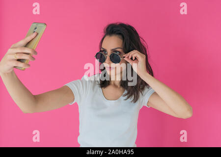 Schöne Mädchen in der Runde Sonnenbrille nimmt eine selfie am Telefon auf einem rosa Hintergrund Stockfoto