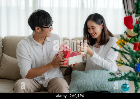 Asiatischer Mann geben die Frau roter Geschenkbox, in der es eine Flasche Parfüm. Die Frau war auf dem Sofa zu Hause abgeholt. Der Weihnachtsbaum in Th Stockfoto