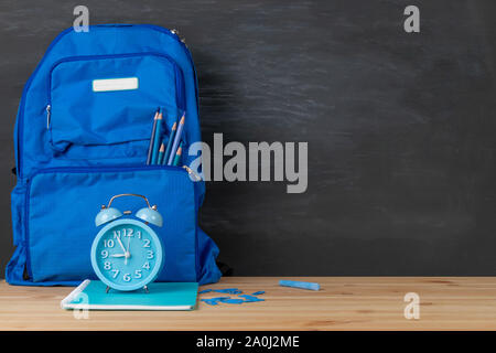 Zurück zu Schule. Rucksack, Wecker und Bücher Himmel blau Ton auf Klassenzimmer Schreibtisch mit schiefertafel Hintergrund. Stockfoto