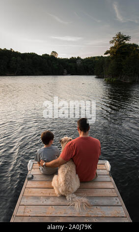 Vater und Sohn sitzen auf Ende eines Docks auf einem See mit ihrem Hund. Stockfoto