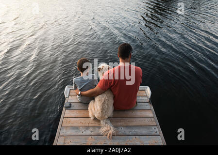 Vater und Sohn sitzen auf Ende eines Docks auf einem See mit ihrem Hund. Stockfoto