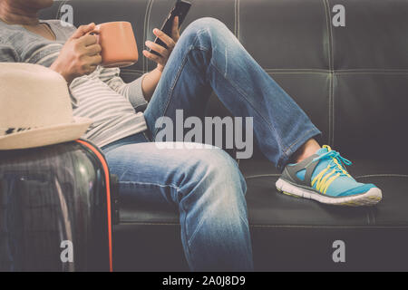 Weibliche Reisende ist entspannend auf leder Sofa holding Handy und Kaffee Tasse mit Reisetasche am Flughafen. Travel Concept. Stockfoto