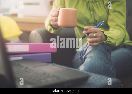 In legerer Kleidung holding Kaffeetasse und pen Geschäftsfrau während der Planung für die Online-bestellung mit Laptop über das Internet. Geschäftliche und Finanzielle. Stockfoto