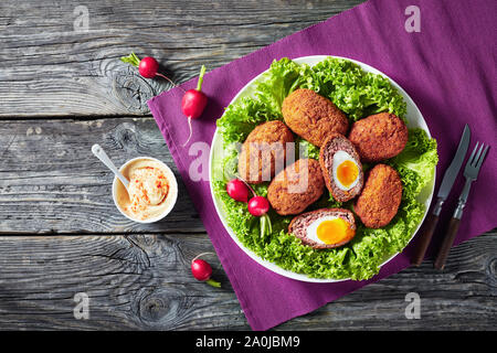 Scotch Eier, weich gekochte Eier mit flüssig Eigelb in Hackfleisch Wurst verpackt, serviert mit Salat und Radieschen auf einem Schild an einem hölzernen Tisch, Blick her Stockfoto