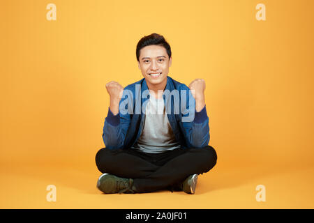 Stattliche fröhlicher junger Asiaten tragen lässige Jeans Denim, Schuhe, Sitzen mit gekreuzten Beinen auf dem Boden und hob die Hände auf. Auf orange backgrou Isoliert Stockfoto