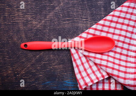 Roter Plastik-Küchenlöffel auf einem schwarzen Tisch mit einem rot karierten Handtuch Stockfoto