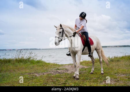 Aktive Frau im Reitsport outfit Bewegen entlang des Flusses auf der Rückseite des Weißen rennpferd Stockfoto