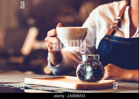 Nahaufnahme des Professional Female barista Hand und halten weiße Tasse Kaffee. Glückliche junge Frau an der Bar im Restaurant Hintergrund. Menschen Stockfoto