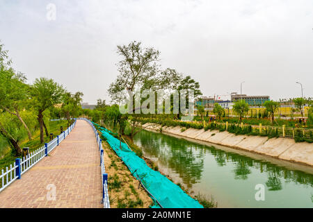 Hotan malerischen Kunlun Lake Park Blick auf einen gepflasterten Gehweg Garten mit Bäumen und einem Wasser Kanal an einem bewölkten Himmel Tag Stockfoto