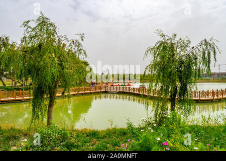 Hotan malerischen Kunlun Lake Park Blick auf einer kurvigen geformten hölzernen Brücke an einem bewölkten Himmel Tag Stockfoto