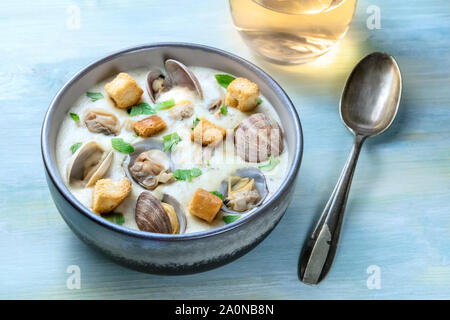 Clam Chowder, dekoriert mit frischer Petersilie und Croutons, auf einem blauen Hintergrund, mit einem Glas Weißwein und einem Löffel Stockfoto