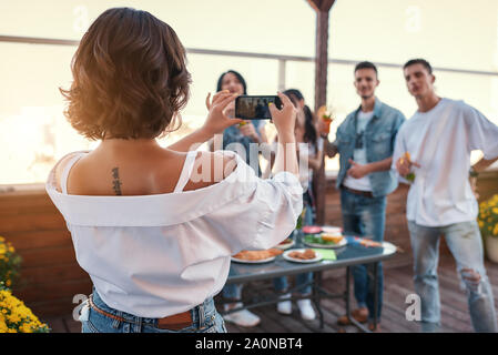 Glückliche Erinnerungen. Rückansicht der jungen Frau, die ein Foto von Ihren Freunden, während mit Party auf dem Dach. Geburtstag Konzept. Freundschaft Stockfoto