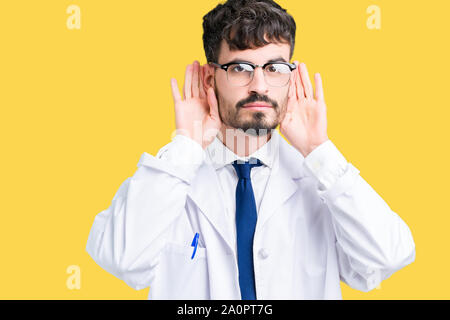 Junge Wissenschaftler Mann mit weißer Mantel über isolierte Hintergrund versuchen, beide Hände am Ohr Geste zu hören, neugierig, für Klatsch und Tratsch. Anhörung pro Stockfoto