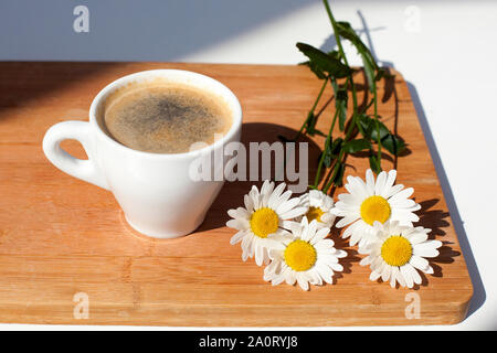 Schwarz Kaffee Tasse mit Schaum und weißen Gänseblümchen Blumen auf einem hölzernen Hintergrund im Sonnenlicht mit Schatten Stockfoto